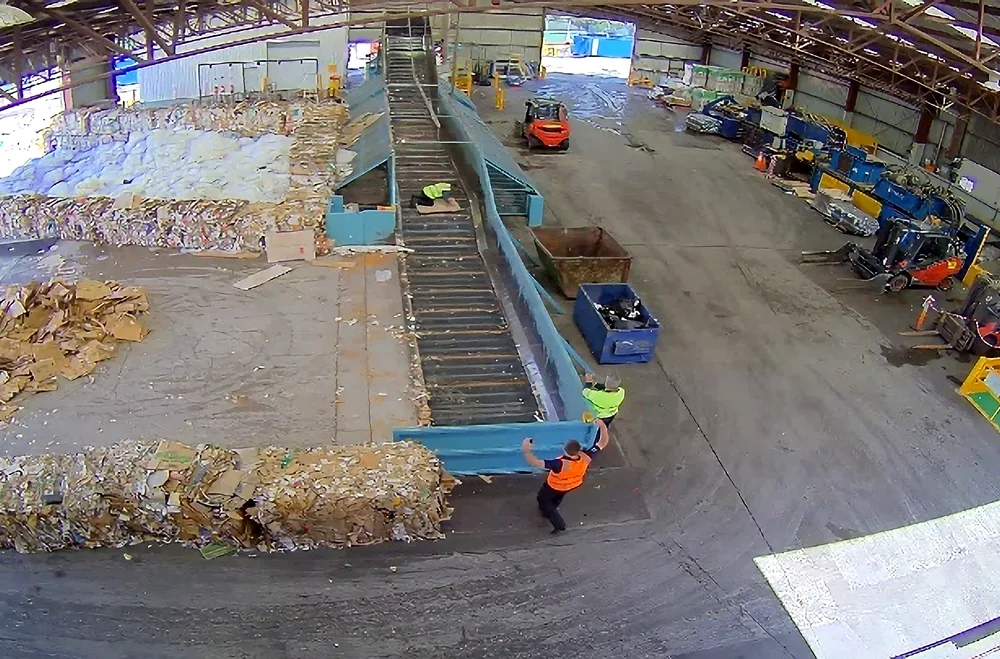 Overhead camera view of a recycling plant: a worker celebrates beside a conveyor as an early WARNY machine integration test works